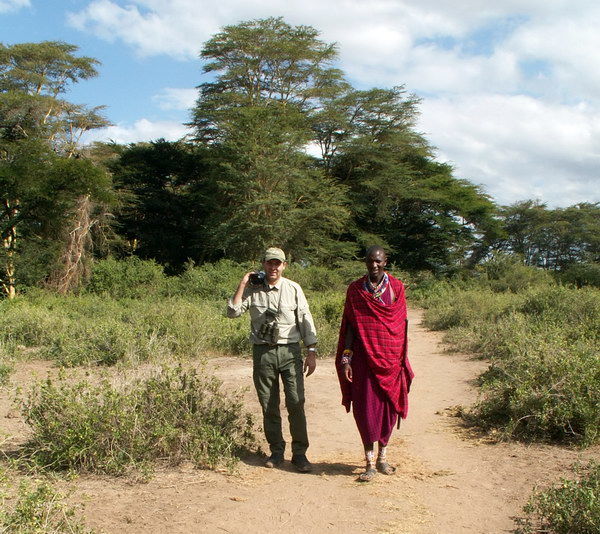 Kameramann Peter Neudecker auf dem Weg zu Fuß in das Masai-Dorf, das im Busch der Masai Mara liegt, in Begleitung eines jugendlichen Morani.
